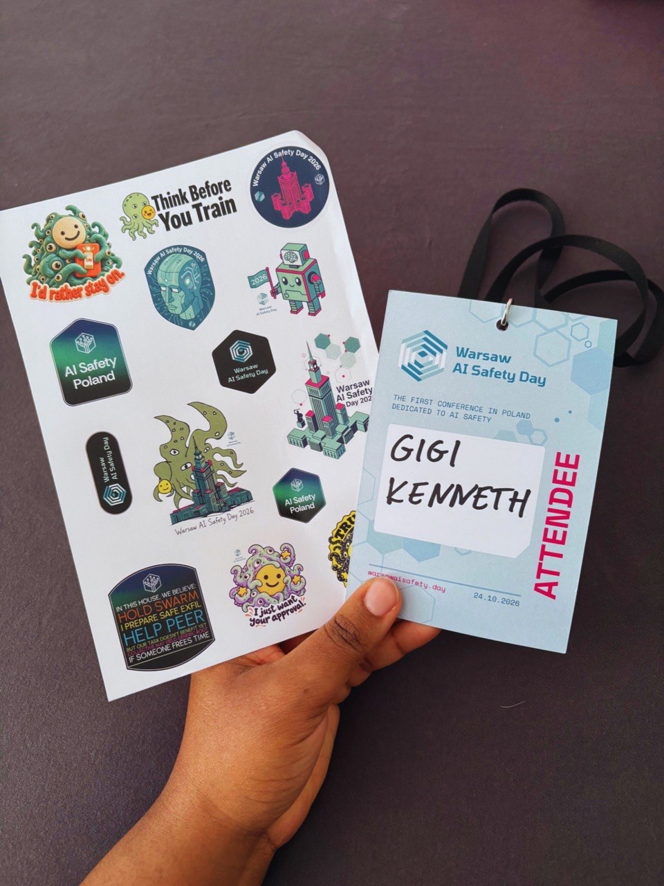 My Warsaw AI Safety Day attendee badge next to a sheet of conference stickers, including a tentacled monster saying I'd rather stay on and an octopus saying Think Before You Train