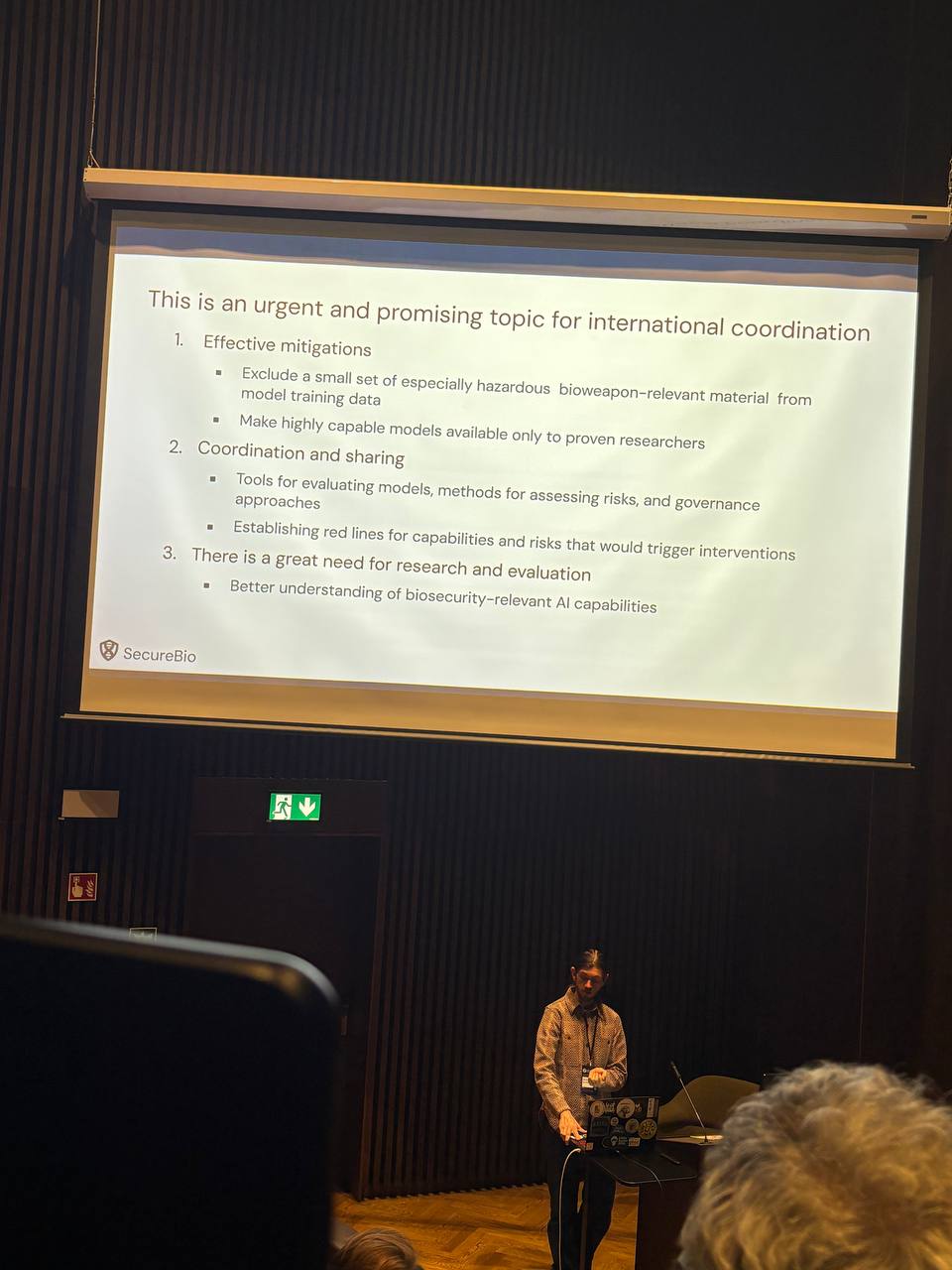 Christopher Stamper of SecureBio presenting a slide titled This is an urgent and promising topic for international coordination, covering mitigations, coordination and sharing, and the need for more research and evaluation