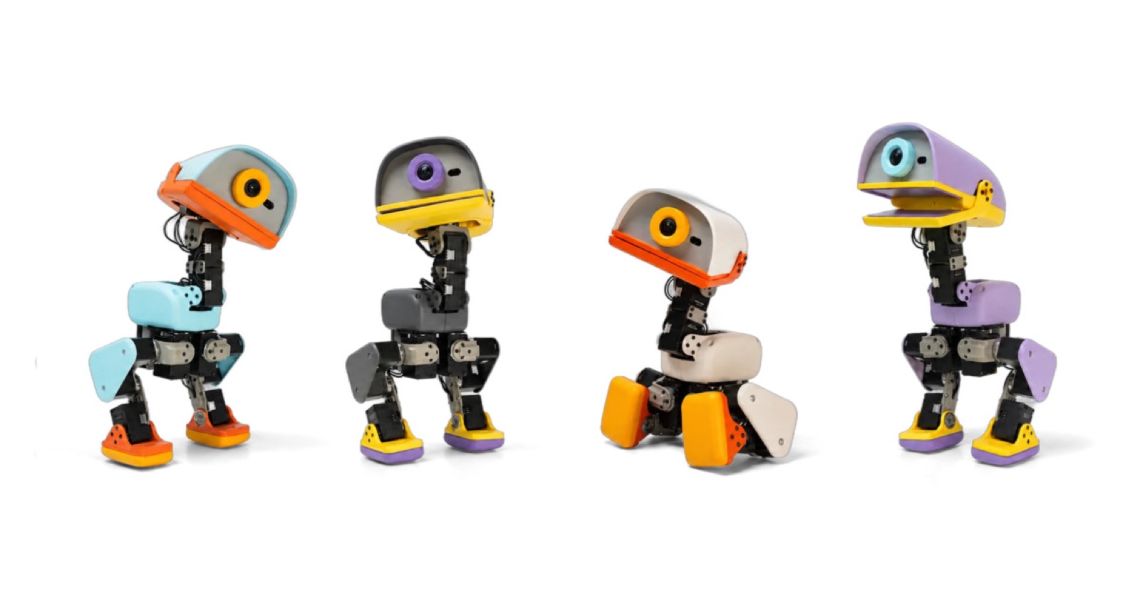 Four small Microduck robots from Pollen Robotics in light blue, grey, white and purple with colourful beaks and feet, standing and sitting in a row