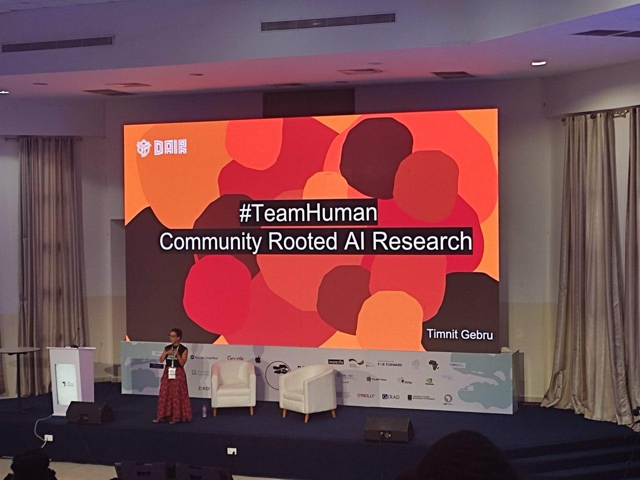 Timnit Gebru on stage at Deep Learning Indaba, slide reading "#TeamHuman: Community Rooted AI Research"